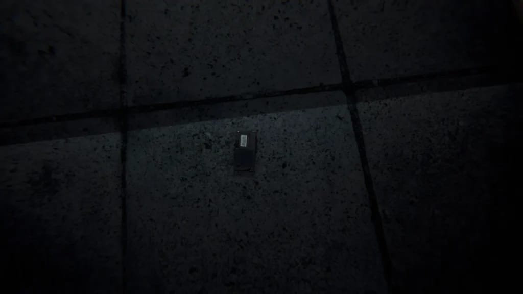 Close-up of a small, dark rectangular electronic device with a barcode on a dim, textured floor in a video game.