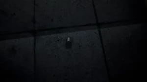 Close-up of a small, dark rectangular electronic device with a barcode on a dim, textured floor in a video game.